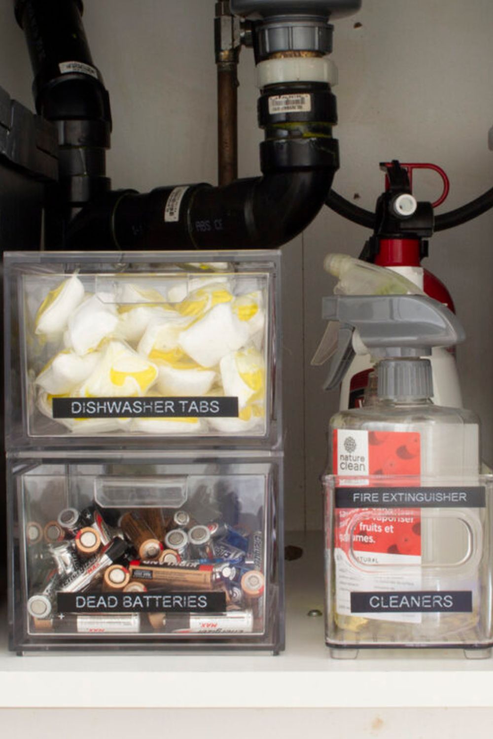 A container of dishwasher tabs on top of a container of dead batteries next to a container of cleaners. The link leads to "How to Organize Under the Kitchen Sink" on HomeMadeLovely.com."